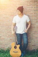 Handsome guy standing holding guitar against the brick wall posing look away. Relaxing holiday. photo