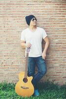 chico guapo de pie sosteniendo la guitarra contra la pared de ladrillo posando mirando hacia otro lado. Vacaciones de relax. foto