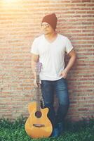 Handsome guy standing holding guitar against the brick wall posing look away. Relaxing holiday. photo