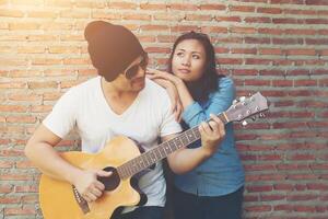 Hipster couple is hugging, looking away and smiling while standing outdoor playing guitar against brick wall, Dating spent great time together. photo
