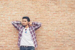 joven apuesto hombre hipster con camisa a cuadros y jeans contra una pared de ladrillos. foto