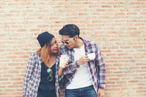 retrato de una joven pareja hipster posando con su novio sosteniendo una taza de café disfrutando de las vacaciones juntos contra el fondo de la pared de ladrillo. foto