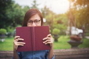 joven hipster sonriendo y cubriendo la cara con su libro rojo mientras se sienta en un banco en el parque. foto