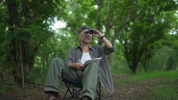 asiatischer senior, pensioniert, sitzt auf einem stuhl und benutzt ein fernglas, das die vögel in den bäumen und im wald betrachtet und die informationen in ein notizbuch aufschreibt. gerne an Urlaubstagen
