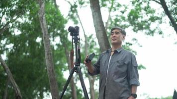 un homme senior asiatique a une caméra dans une forêt et un itinéraire d'examen. vieil homme regardant dans des jumelles en bois tout en se promenant en plein air. touriste masculin explorant le terrain. concept de tourisme et de randonnée. video