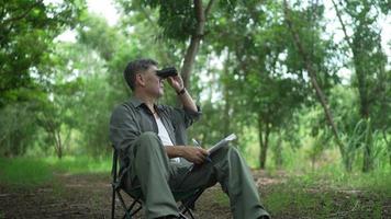asiatisk senior man, pensionerad, som sitter på en stol använder en kikare och tittar på fåglarna i träden, i skogen och skriver ner informationen i en anteckningsbok. gärna på semesterdagar video