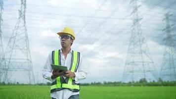 Font View.Electrical engineer wearing a Yellow helmet and safety carrying using tablet vest walking near high voltage electrical lines towards power station on the field.