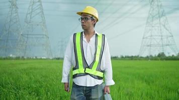 fonte view.engenheiro elétrico usando um capacete amarelo e colete de caixa de ferramentas de transporte de segurança andando perto de linhas elétricas de alta tensão em direção à usina no campo. video