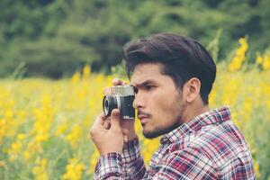 joven apuesto hombre hipster fotografiando con cámara retro con campo de flores amarillas de la naturaleza. foto