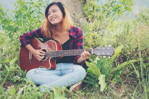 joven hermosa mujer hipster tocando la guitarra sentada en el césped en el parque, relajándose con la naturaleza. foto