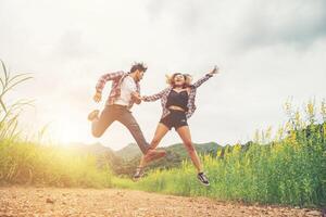 joven hipster pareja enamorada saltando al aire libre en el campo de flores amarillas con fondo de puesta de sol de montaña. salta alto disfrutando de la libertad y disfruta. foto