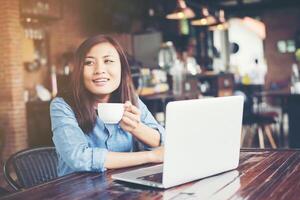 hermosa joven hipster sentada en una cafetería, sosteniendo una taza de café, relájese y juegue con su computadora portátil, mirando hacia otro lado, feliz y divertida. concepto de estilo de vida. foto