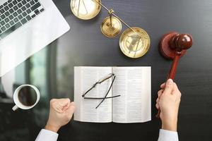 justice and law concept.Top view of Male judge hand in a courtroom with the gavel and brass scale and computer and open bible book on dark wood table photo