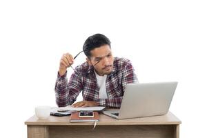 Thoughtful young man holding glasses looking at laptop while sitting at his working place. photo