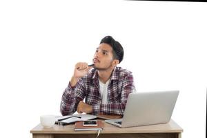 Thoughtful young man holding hand on chin and looking away while sitting at his working place isolated on white background.