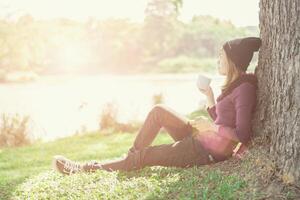 Smiling woman with cup coffee enjoying near riverside against a tree in morning sunlight photo