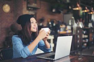 Beautiful young hipster woman sitting in a coffee shop, holding a cup of coffee relax and play with her laptop, looking away, happy and fun. Lifestyle concept.
