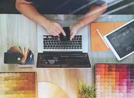 vista superior de la mano del diseñador de interiores usando un teléfono inteligente y eligiendo una muestra de alfombra con una nueva computadora portátil moderna en blanco y una tableta digital con tablero de material en un escritorio de madera como concepto foto