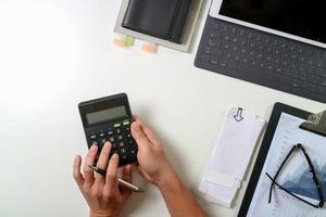 vista superior de la mano de hombre de negocios que trabaja con finanzas sobre costos y calculadora y portátil con teléfono móvil en el escritorio en la oficina moderna foto