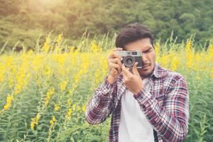 Young handsome hipster man photographing with retro camera with nature yellow flower field. photo