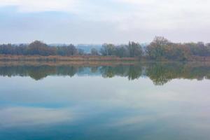 Cold foggy river in the morning with overgrown banks and displaying clouds in the water. photo