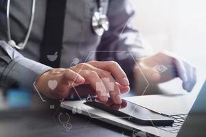 close up of smart medical doctor working with mobile phone and laptop computer and stethoscope on dark wooden desk photo