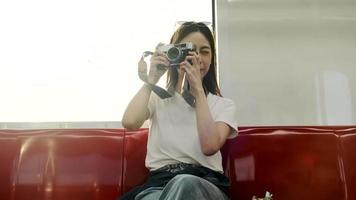 una hermosa turista asiática con cámara se sienta en un asiento rojo, viaja en tren aéreo, toma una foto instantánea cuando se transporta en una vista urbana, el estilo de vida de los pasajeros de la ciudad en tren, feliz viaje de vacaciones. video