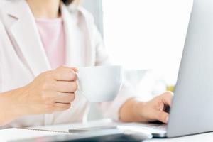 primer plano de una mujer de negocios usando una computadora portátil y sentada en el escritorio de la oficina, sosteniendo una taza de café. foto