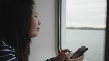 asiatische frau, die smartphone benutzt und durch das fenster auf dem boot schaut, bildschirm auf dem smartphone verschiebt. Sitzen auf dem Boot am Fenster mit wunderschönem Meerblick draußen