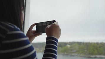 cierra a una mujer asiática tomando una foto a través de la ventana del barco, usando un smartphone para tomar una foto. sentado en el barco junto a la ventana con hermosas vistas al mar afuera video