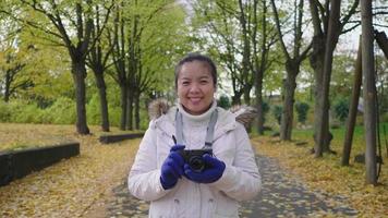 Asian woman holding camera standing and smiling on the road in the park. Autumn outfits, Good weather, yellow leaves falling on the street. A lot of tree background, Stockholm, Sweden in Autumn video