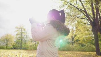 mulher asiática tirando uma foto de vista no parque no outono. roupas de outono, tempo bom, folhas amarelas caindo na rua. muita árvore e fundo de luz solar, estocolmo, suécia no conceito de outono video