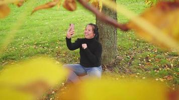 glad asiatisk kvinna som sitter och tar ett videosamtal på smartphone under trädet i parken på hösten, kvinna klädd i svart långärmad skjorta, gult blad på trädet, sverige, tittar genom trädet video