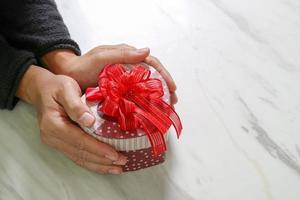 entrega de regalos, mano de hombre sosteniendo una caja de regalo en forma de corazón en un gesto de dar sobre fondo de mesa de mármol gris blanco foto