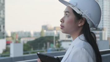 een zelfverzekerde vrouwelijke architect in een witte hoed kijkt met een glimlach naar de camera. vrouwelijke bouwingenieur met een tabletcomputer op een bouwplaats. video