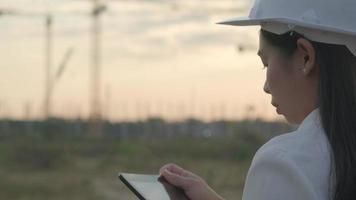 engenheiro de construção feminino com um computador tablet em um canteiro de obras ao pôr do sol. arquiteto de mulher confiante no capacete branco, olhando para um canteiro de obras. video