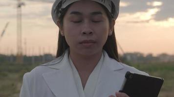 una arquitecta segura de sí misma con un sombrero blanco mira a la cámara con una sonrisa. ingeniera de construcción con una tableta en un sitio de construcción. video