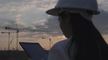 Female construction engineer with a tablet computer at a construction site at sunset. Confident woman architect in white helmet looking at a construction site.