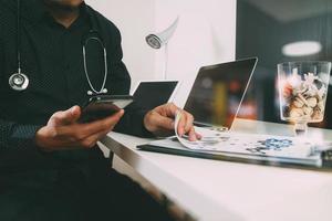 médico inteligente que trabaja con teléfonos inteligentes y tabletas digitales y computadoras portátiles y estetoscopio en un escritorio de madera en una oficina moderna foto