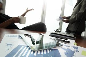 business documents on office table with smart phone and digital tablet and stylus and two colleagues discussing data in the background photo