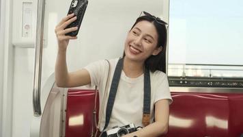 Young beautiful Asian female tourist online chatting, mobile phone video call, check-in, and social media posting, travel urban train transport, city passenger lifestyle, happy journey vacation trip.