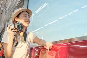 Beautiful Asian female tourist sits in a red seat, traveling by train, taking snapshot photo, transporting in suburb view, enjoy passenger lifestyle by railway, happy journey vacation. photo