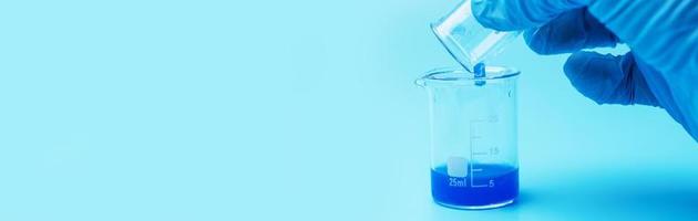 Close-up on the hands of a scientist wearing blue rubber gloves and pouring liquid chemicals from glass to glass. photo