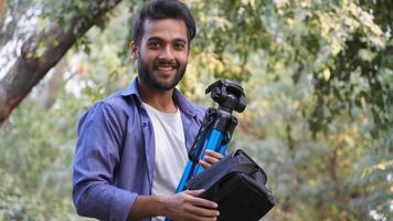 Professional photographer. Portrait of confident young man in shirt holding hand on camera on tripod photo