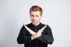 Portrait of a young man in a black shirt shows a prohibition sign with his hands on a white background. Copy, empty space for text photo