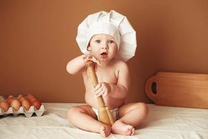 Little baby boy in a chef's hat sitting on the table holds a wooden rolling pin on a brown background photo