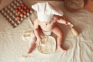 Little boy baker in a chef's hat sitting on the table playing with flour on a brown background with a wooden rolling pin, a round rustic sieve and eggs. Top view. Copy, empty space for text photo
