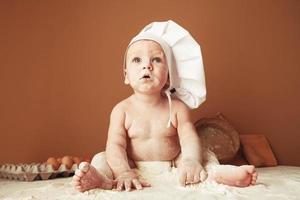 panadero de niño con sombrero de chef sentado en la mesa jugando con harina sobre un fondo marrón con un rodillo de madera, un tamiz rústico redondo y huevos. copiar, espacio vacío para texto foto