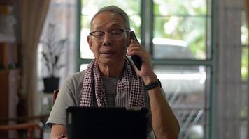 Elderly man talking on mobile phone while working on the desk with digital tablet at home. video