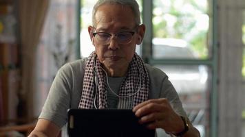 älterer mann, der zu hause ein digitales tablet mit kopfhörern auf dem schreibtisch verwendet. video